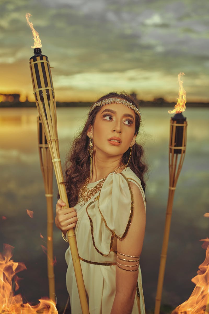 A stunning woman in a Greek-style dress holding a torch near a lake during sunset.